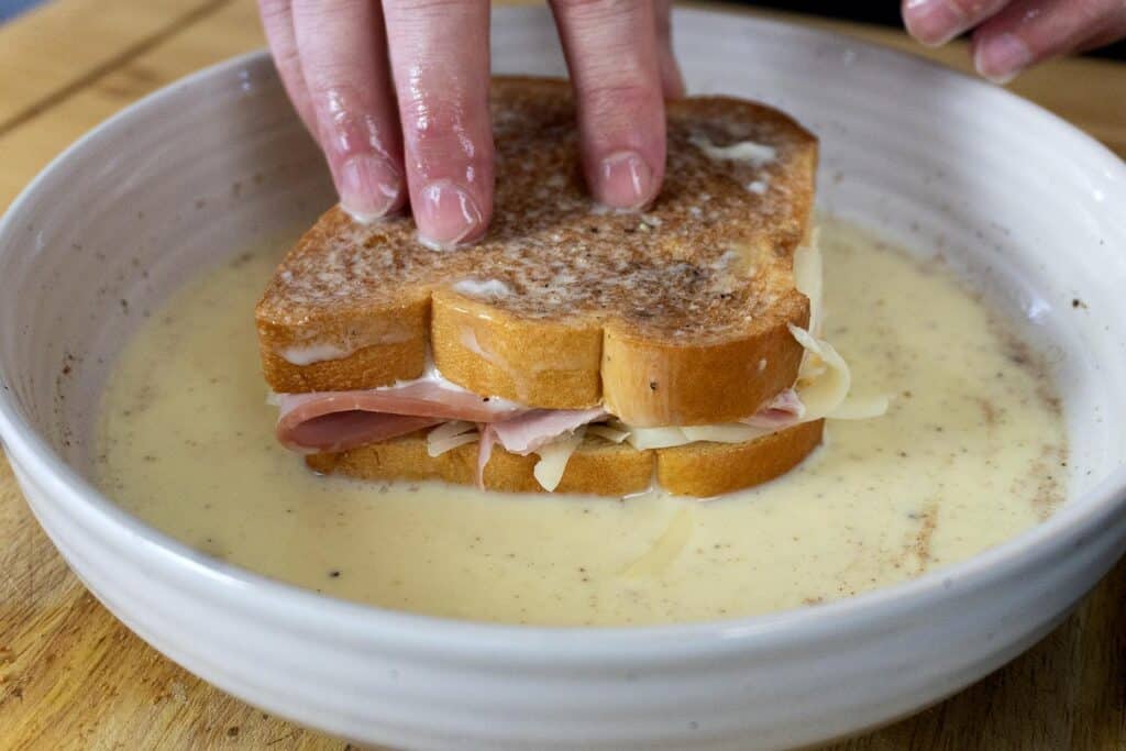 A person presses a ham and cheese sandwich into a shallow bowl filled with an egg and milk mixture, preparing it for cooking.