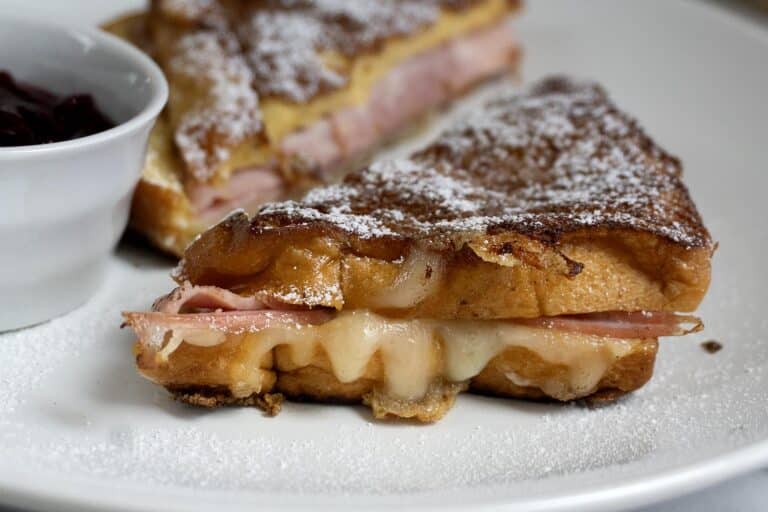 A close-up of a Monte Cristo sandwich with ham and melted cheese on toasted bread, dusted with powdered sugar, served on a white plate with a small dish of red jam.