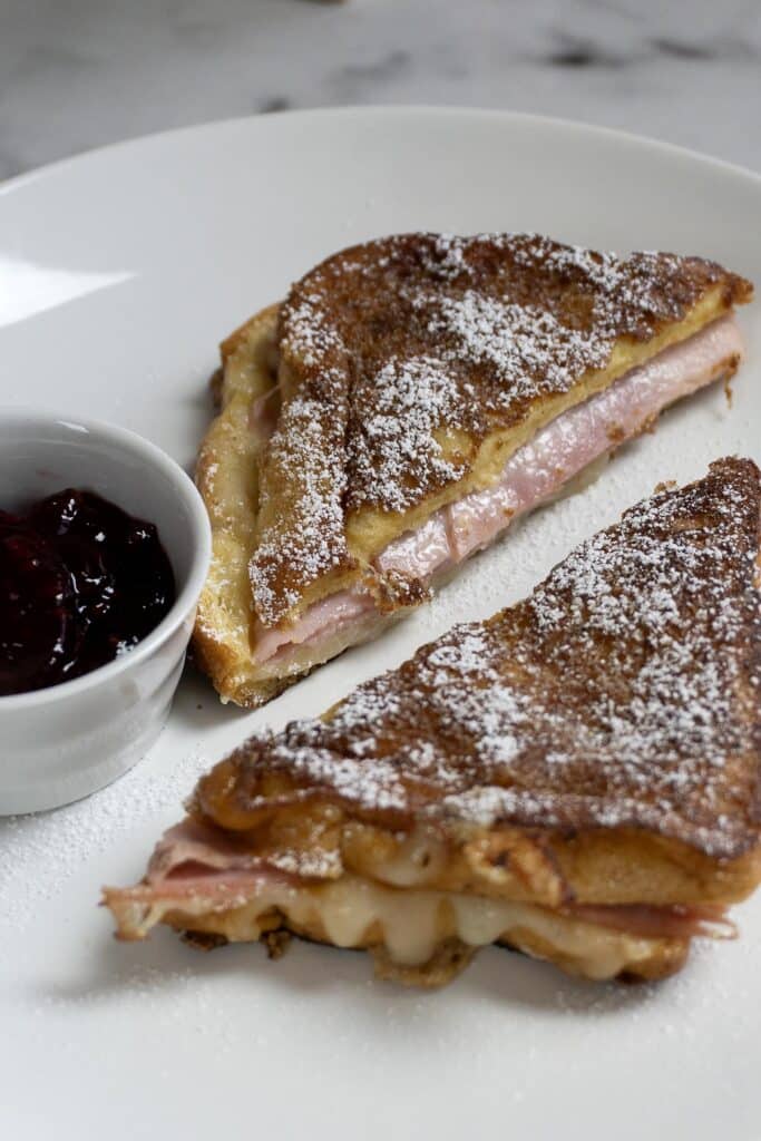 A plate with two halves of a ham and cheese sandwich dusted with powdered sugar, next to a small white cup filled with dark fruit jam. The food is served on a white plate with a marble surface underneath.