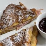 Two powdered sugar-dusted sandwich halves filled with ham are on a white plate next to a small bowl of dark berry jam.