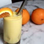 A glass of orange smoothie with a metal straw and an orange slice garnish sits on a marble table, with two whole oranges in the background.