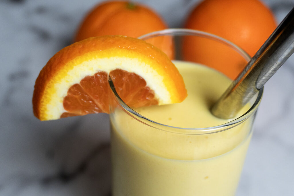 A glass of orange smoothie garnished with a fresh orange slice and a metal straw, with two whole oranges in the background on a marble surface.