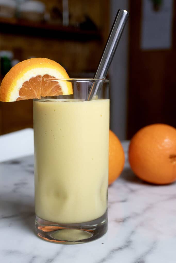 A glass of creamy orange smoothie with ice, garnished with an orange slice and a metal straw, sits on a marble surface. Two whole oranges are in the background.
