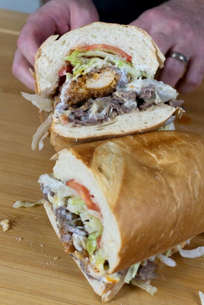 A close-up of a sandwich cut in half, filled with fried chicken, roast beef, lettuce, tomato, and mayonnaise, resting on a wooden cutting board with a persons hands holding the sandwich.