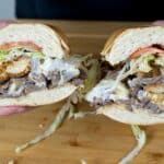 A person holds a large sandwich cut in half, filled with sliced steak, fried chicken, lettuce, tomato, and melted cheese, with mayonnaise spilling out, over a wooden surface.
