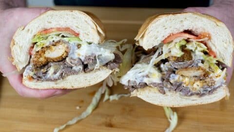 A person holds a large sandwich cut in half, filled with sliced steak, fried chicken, lettuce, tomato, and melted cheese, with mayonnaise spilling out, over a wooden surface.