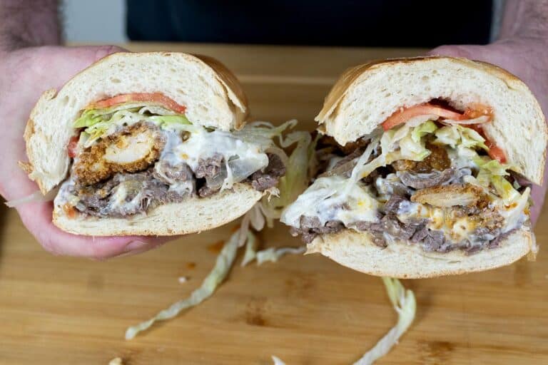 A person holds a large sandwich cut in half, filled with sliced steak, fried chicken, lettuce, tomato, and melted cheese, with mayonnaise spilling out, over a wooden surface.