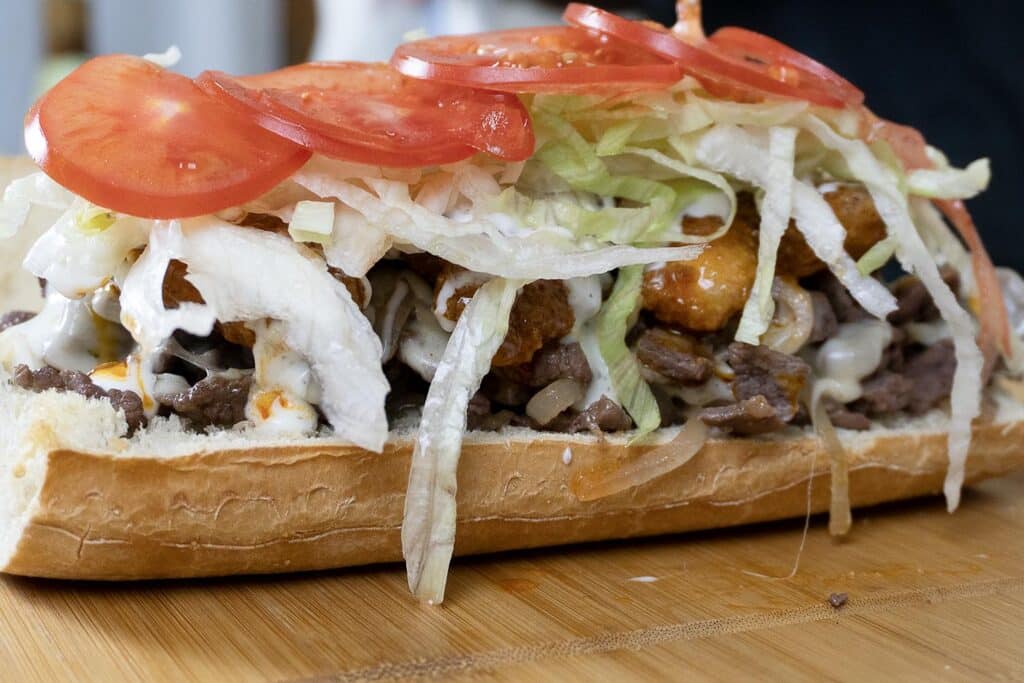 A close-up of a sub sandwich on a wooden board, filled with sliced steak, lettuce, onions, tomato slices, fried bits, and drizzled with a creamy sauce.