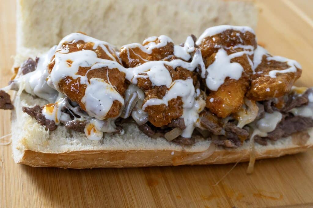 A sandwich on a hoagie roll filled with sliced steak, melted cheese, fried chicken pieces coated in sauce, and drizzled with ranch dressing, served on a wooden surface.