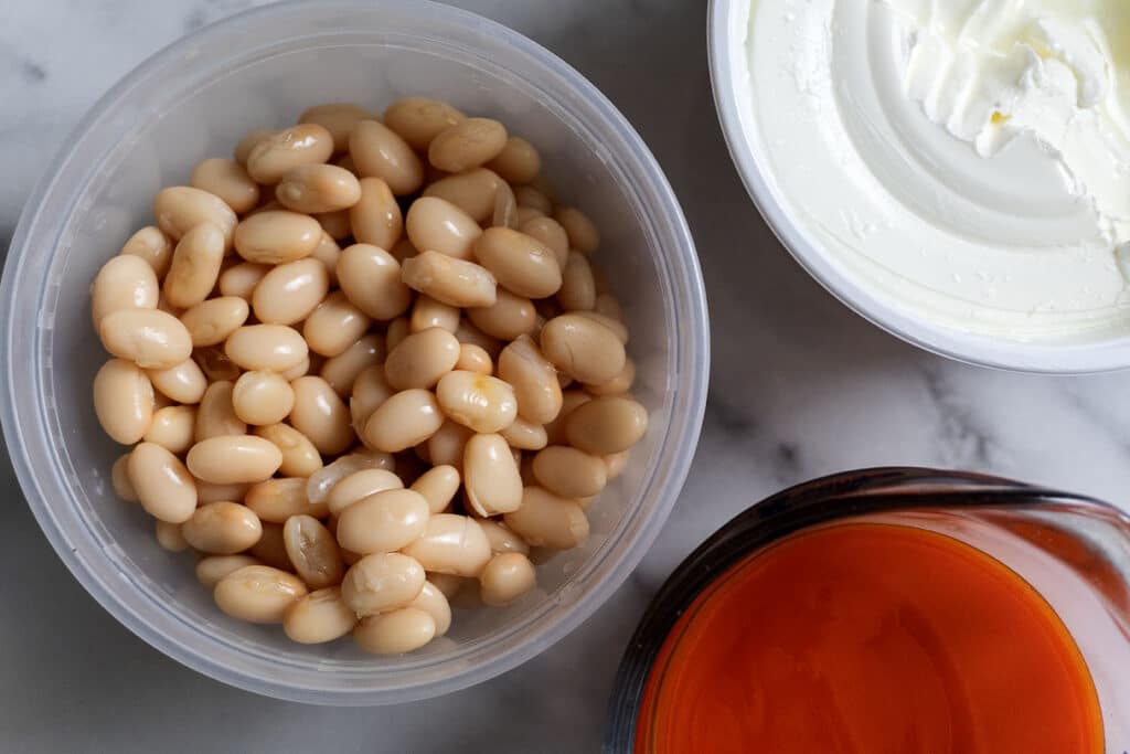 A plastic container of white beans, a bowl of creamy white cheese or yogurt, and a glass measuring cup filled with bright red sauce are arranged on a marble surface.