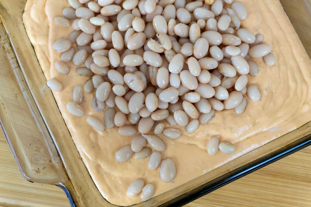 A glass baking dish filled with a creamy orange mixture, topped with a pile of white beans, on a wooden surface.