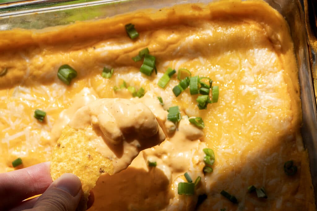 A hand holds a tortilla chip dipped in creamy, cheesy baked dip topped with sliced green onions in a glass baking dish.