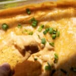 A hand holds a tortilla chip dipped in creamy, cheesy baked dip topped with sliced green onions in a glass baking dish.