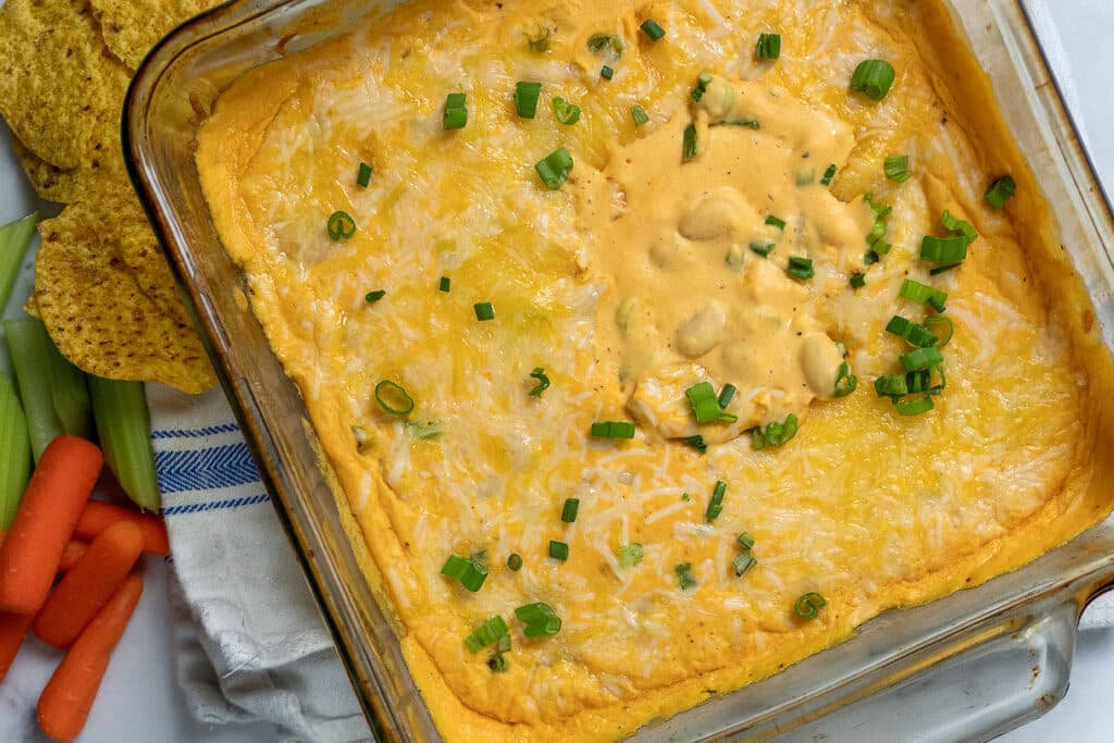 A glass baking dish filled with cheesy buffalo chicken dip, topped with melted cheese and chopped green onions. Tortilla chips, celery sticks, and baby carrots are arranged next to the dish on a white towel.