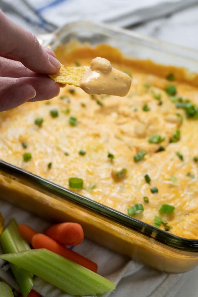 A hand holds a tortilla chip dipped in creamy, orange-colored dip from a glass baking dish topped with chopped green onions. Celery sticks and baby carrots are visible in the foreground.