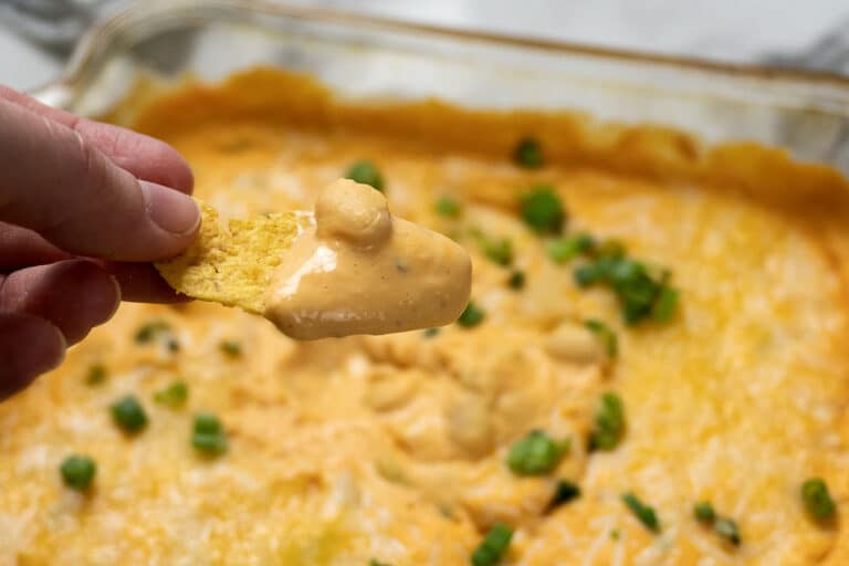 A hand holds a tortilla chip dipped in creamy, orange cheese dip above a baking dish filled with the same dip, topped with chopped green onions.