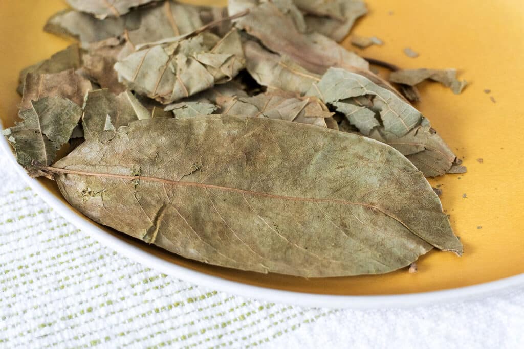 A close-up of several dried bay leaves on a yellow plate, with a textured white cloth underneath.