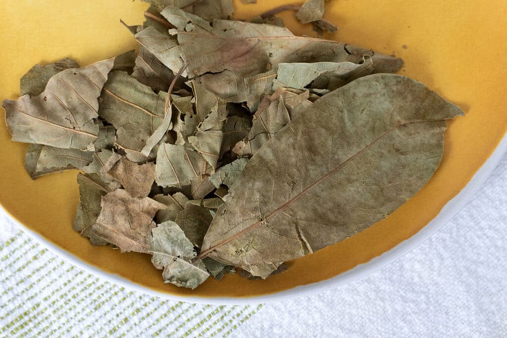 A close-up of dried bay leaves on a yellow plate, placed on a white surface with a green-striped cloth partially visible underneath.