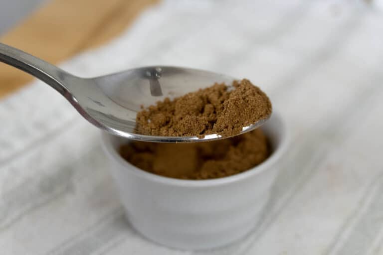A close-up of a spoon holding ground spice over a small white ramekin filled with the same brown powder, placed on a white textured surface.