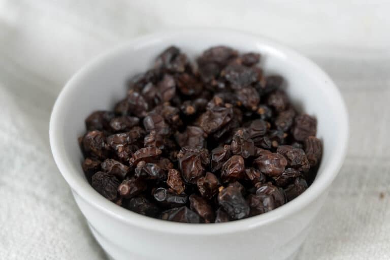 A white bowl filled with dark, dried currants sits on a textured, light-colored cloth background.