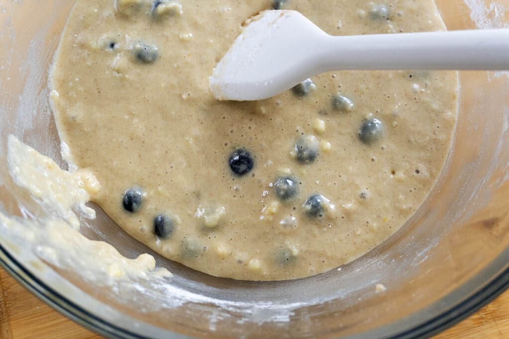 A mixing bowl filled with thick batter containing blueberries, being stirred with a white spatula. The bowl sits on a wooden surface.