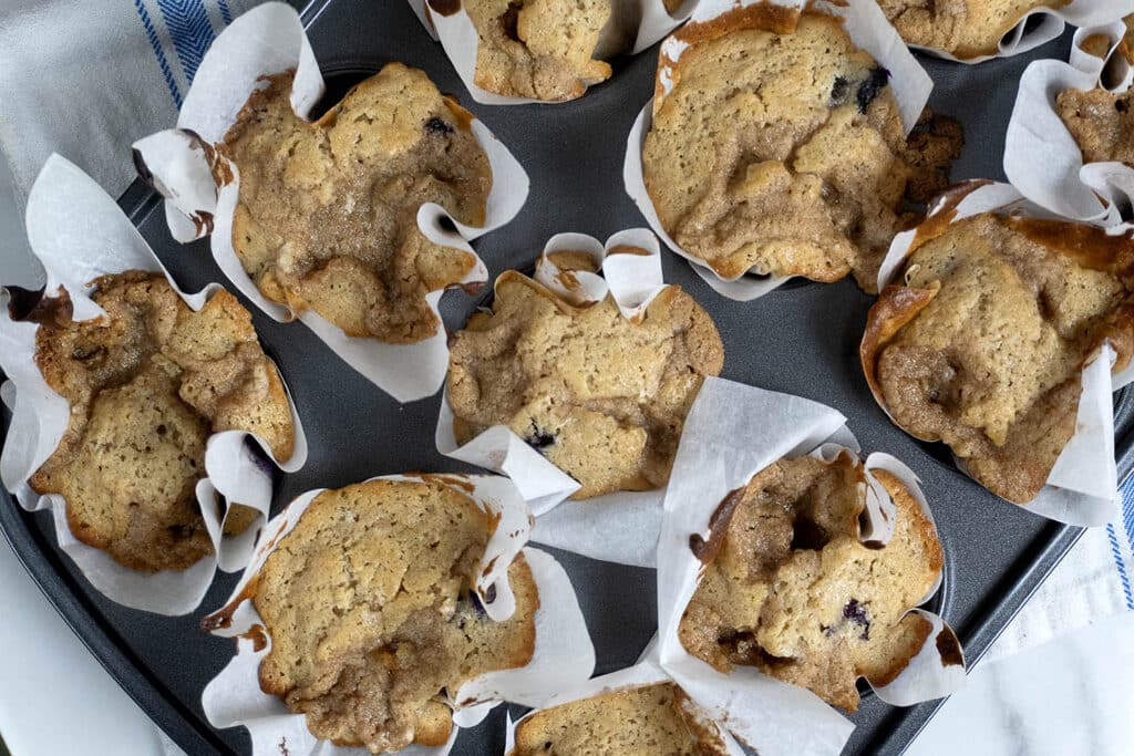 A close-up of several freshly baked muffins in a muffin tin, each wrapped in white parchment paper. The muffins are golden brown with slightly uneven tops and visible blueberries.