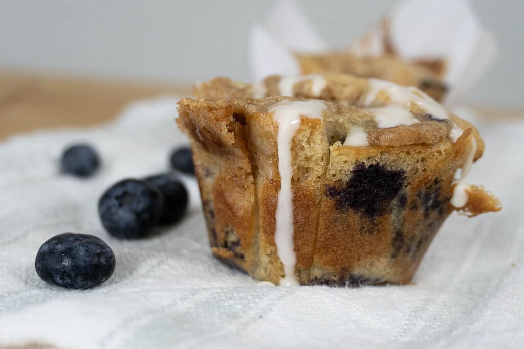 A close-up of a blueberry muffin drizzled with white icing, sitting on a textured white cloth, with fresh blueberries scattered nearby.