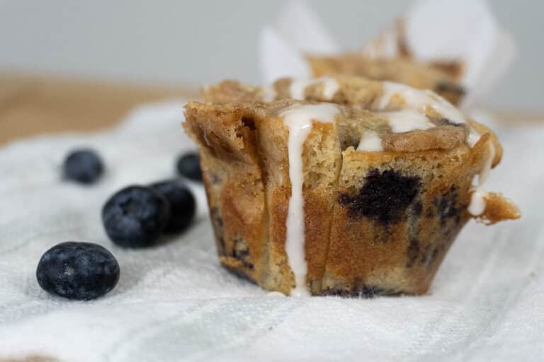 A close-up of a blueberry muffin drizzled with white icing, sitting on a textured white cloth, with fresh blueberries scattered nearby.