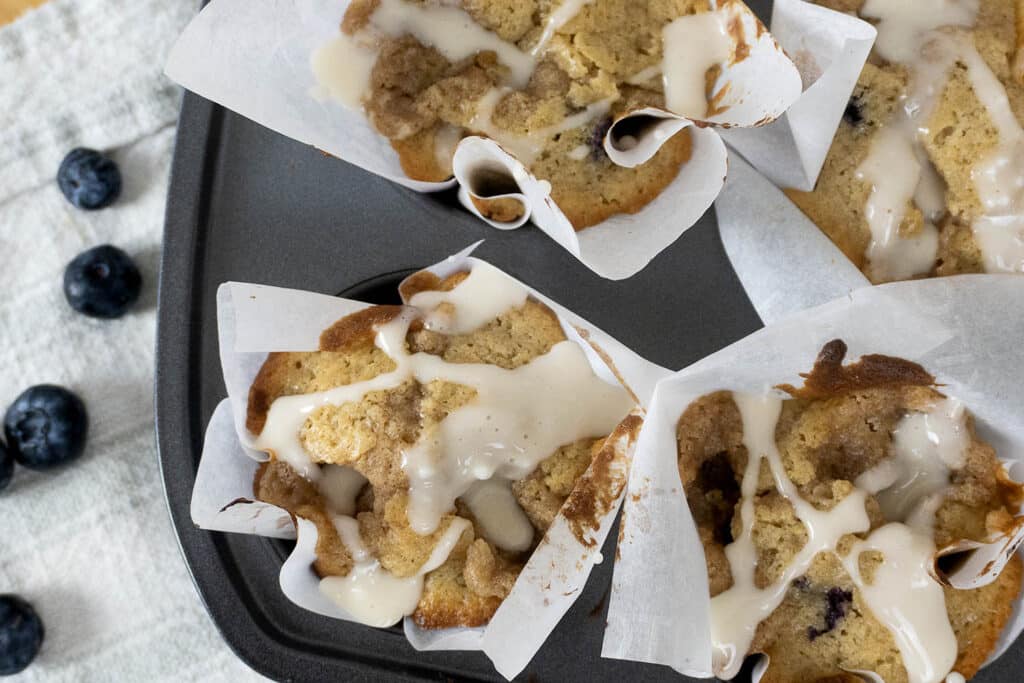 Four blueberry muffins with crumb topping and white glaze sit in parchment paper liners in a muffin tin. A few fresh blueberries are scattered on a white cloth beside the tin.