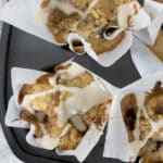 Four blueberry muffins with crumb topping and white glaze sit in parchment paper liners in a muffin tin. A few fresh blueberries are scattered on a white cloth beside the tin.