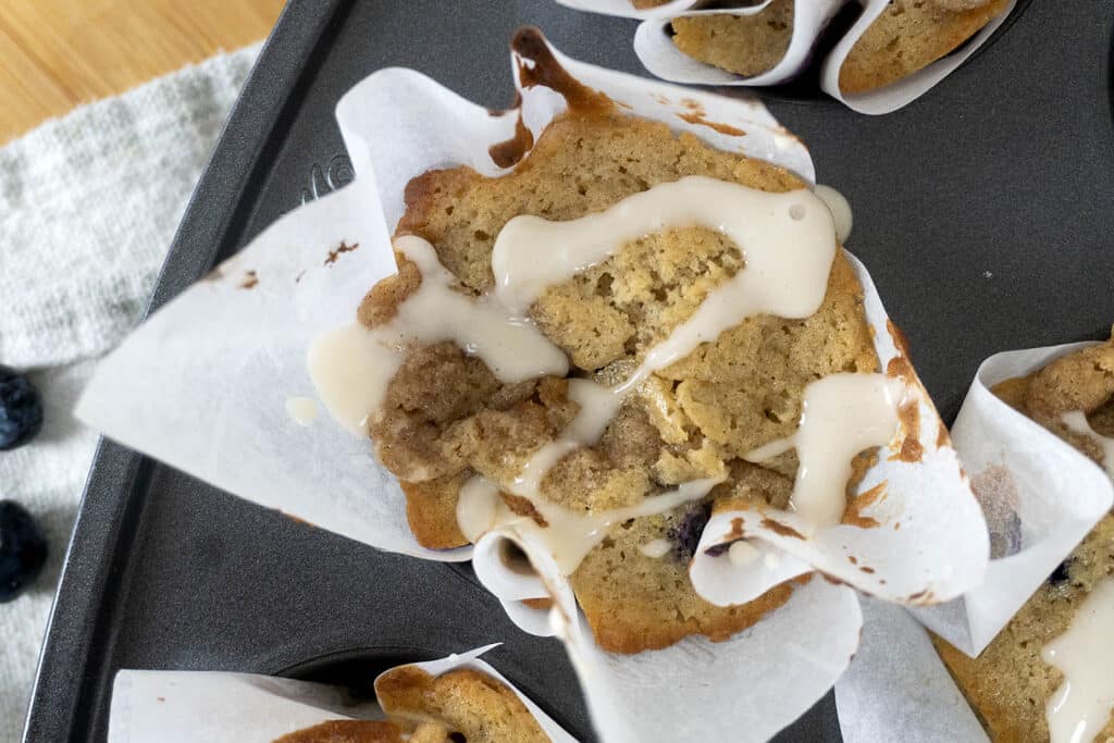 A close-up of a muffin in a white paper liner, drizzled with glaze, sitting in a muffin tin. The muffin has a golden-brown top with visible cracks and a slightly uneven surface.