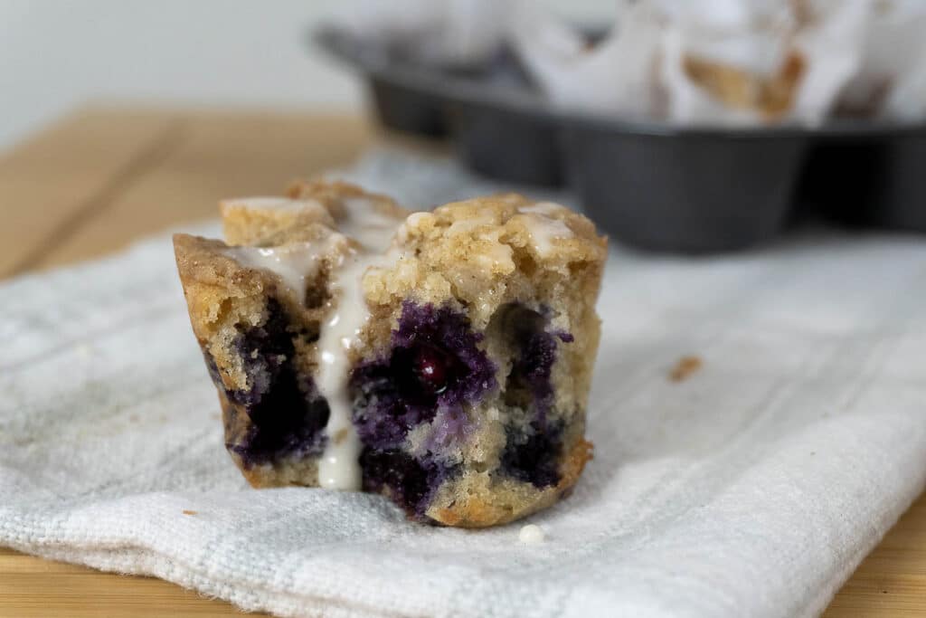 A partially eaten blueberry muffin with icing drips down the side, sitting on a cloth napkin. In the background, more muffins are in a dark muffin tin.