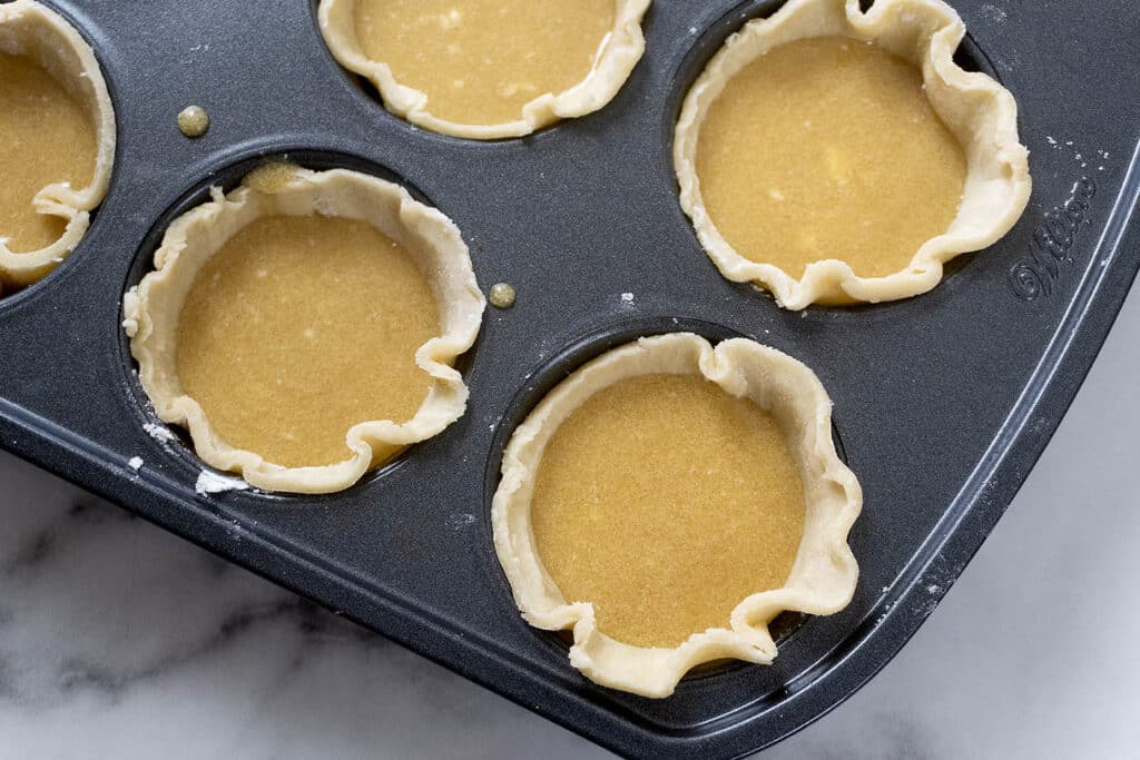 A muffin tin filled with unbaked mini pie crusts, each holding a smooth yellow filling, sits on a marble countertop.
