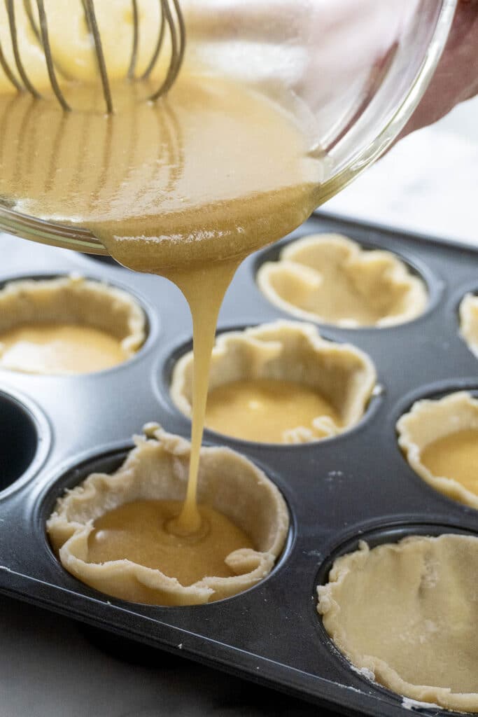 A close-up of batter being poured from a bowl into unbaked pie crusts in a muffin tin, preparing mini pies or tarts. A whisk is visible in the bowl.