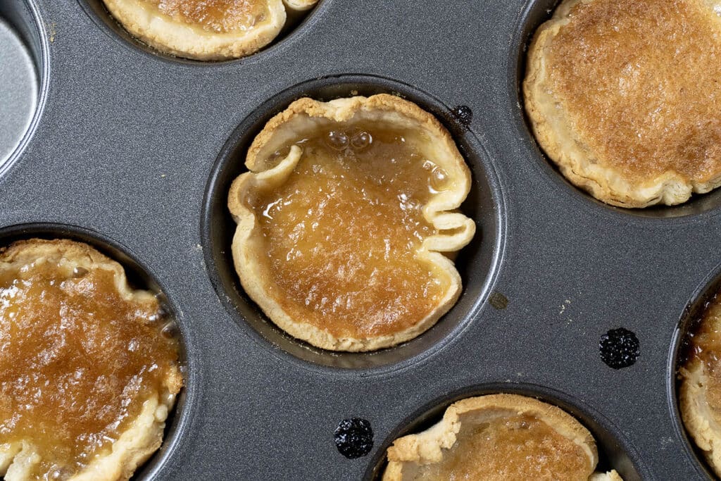 Close-up of a muffin tin holding several golden brown butter tarts, with flaky pastry crusts and caramelized filling, some filling slightly overflowing onto the tin.