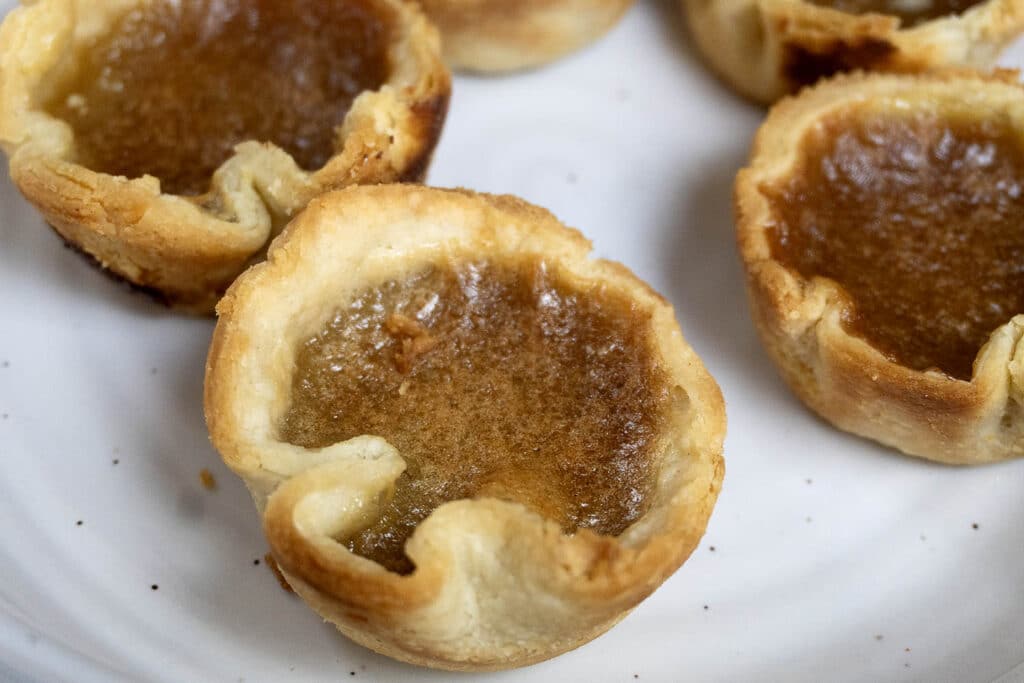 Close-up of several golden brown butter tarts with flaky pastry crusts and gooey filling, displayed on a white plate.