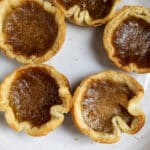 Five butter tarts with golden, flaky pastry crusts and caramelized, glossy filling are arranged on a white plate.