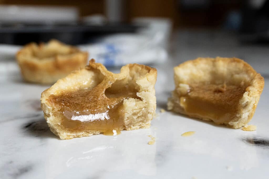 Two butter tarts are shown on a marble surface, one cut in half to reveal a gooey, golden caramel filling inside a flaky pastry shell.
