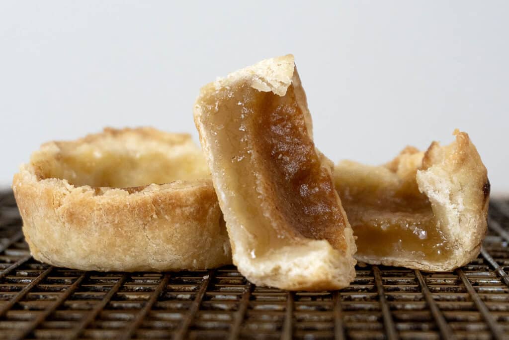 A close-up of a butter tart cut in half, revealing its gooey, golden filling and flaky pastry crust, sitting on a cooling rack against a plain background.
