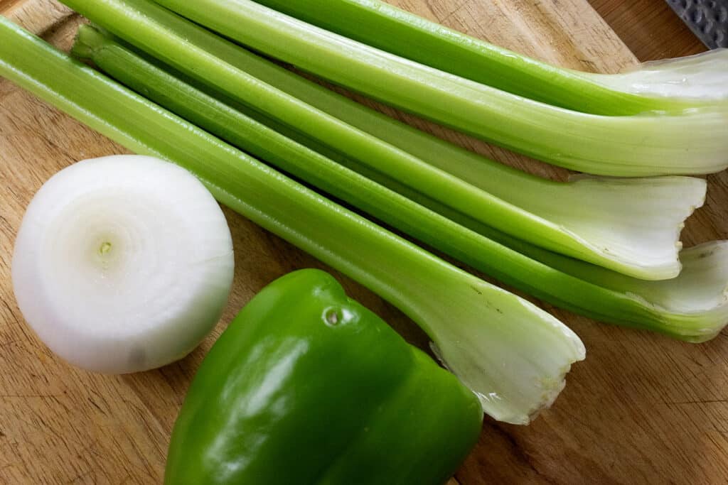 Three stalks of celery, a whole white onion, and a green bell pepper sit on a wooden cutting board, ready for preparation.