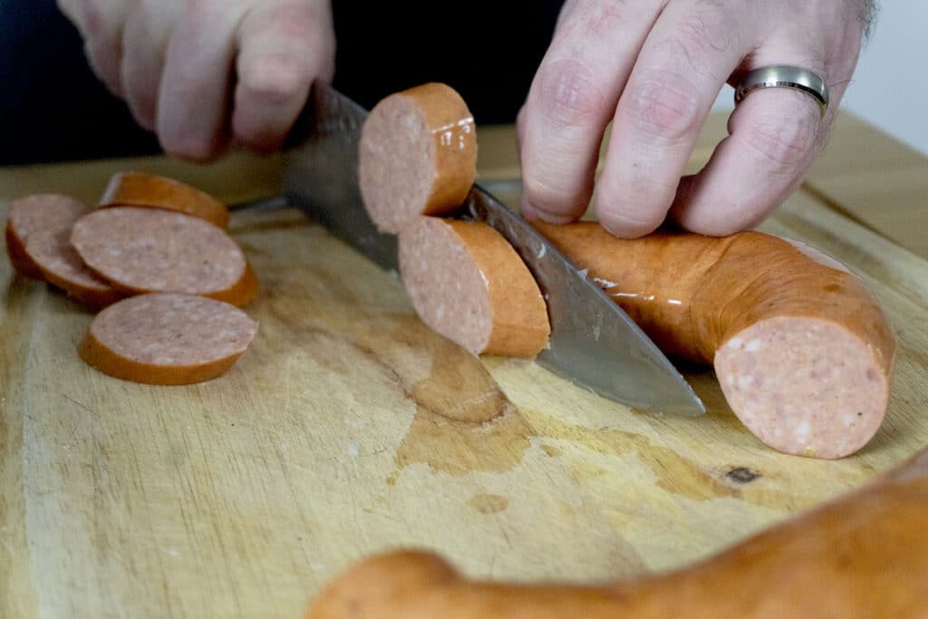 A person wearing a ring slices a sausage into thick pieces on a wooden cutting board with a large kitchen knife.