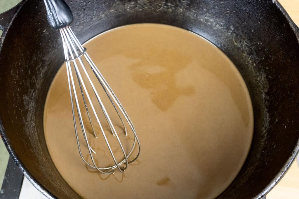 A metal whisk rests in a black pot filled with smooth, brown gravy or sauce.