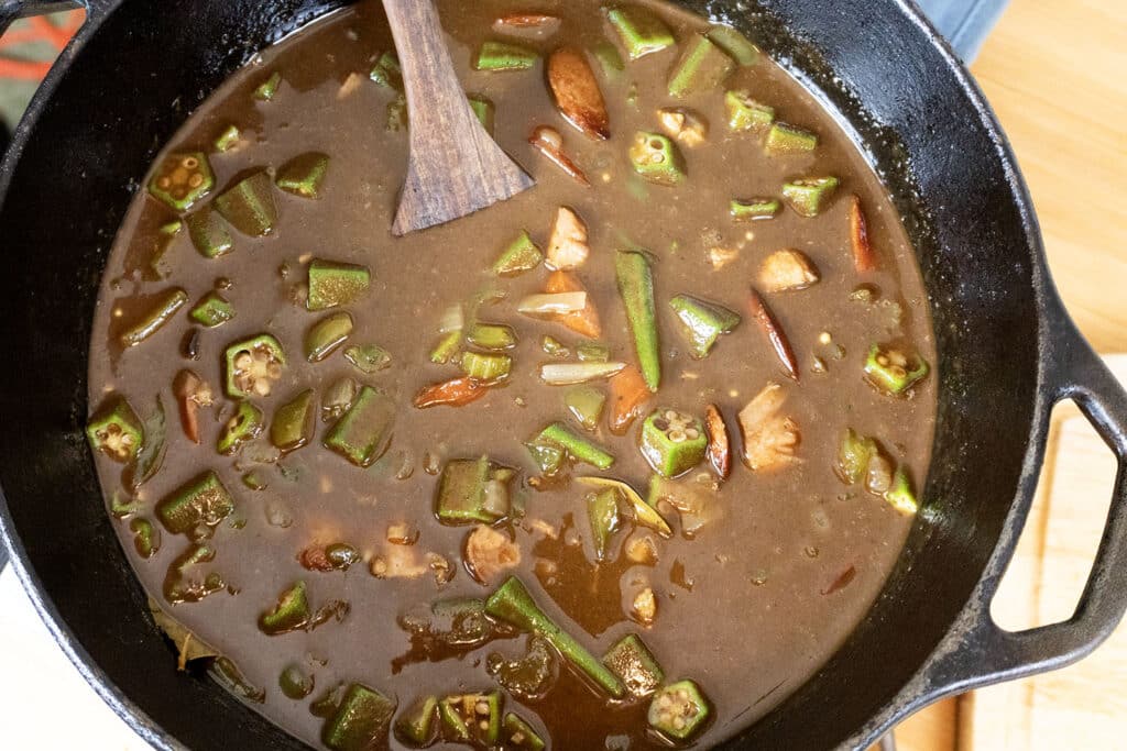 A pot of gumbo with chopped okra, tomatoes, and other vegetables in a brown broth, being stirred with a wooden spoon.