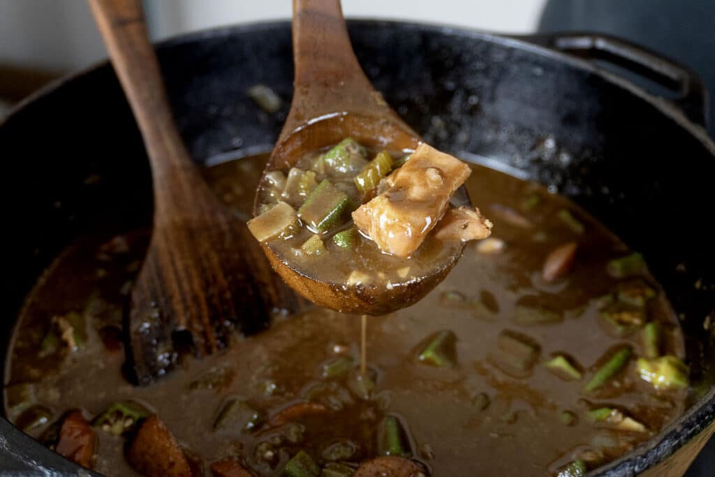 A wooden spoon lifts a portion of gumbo with okra and meat from a pot filled with the hearty, brown stew. Another wooden utensil rests inside the pot.