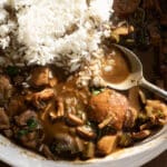 A bowl of gumbo with chunks of meat, sausage, and vegetables in a dark brown sauce, served with white rice. A spoon rests in the bowl.