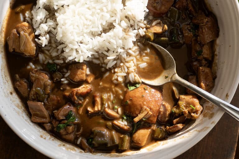 A bowl of gumbo with chunks of meat, sausage, and vegetables in a dark brown sauce, served with white rice. A spoon rests in the bowl.