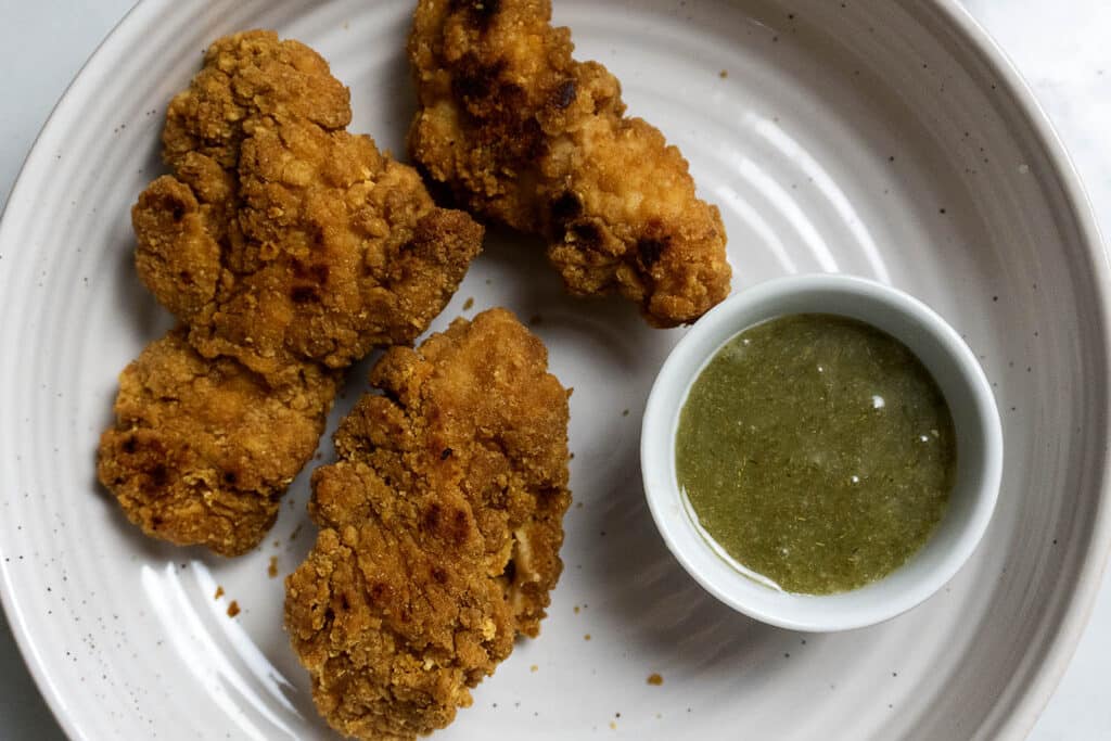 Three pieces of crispy fried chicken are served on a white plate with a small bowl of green dipping sauce on the side.