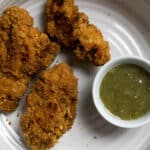 Three pieces of crispy fried chicken are served on a white plate with a small bowl of green dipping sauce on the side.