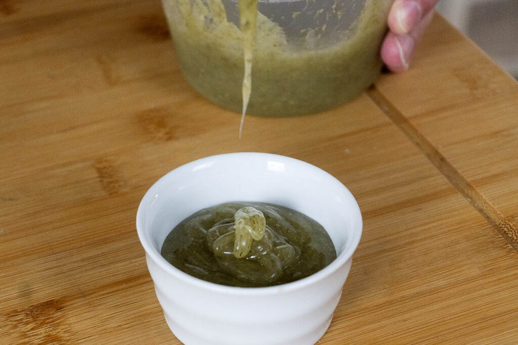 A thick green sauce is being poured from a container into a small white ramekin on a wooden surface.