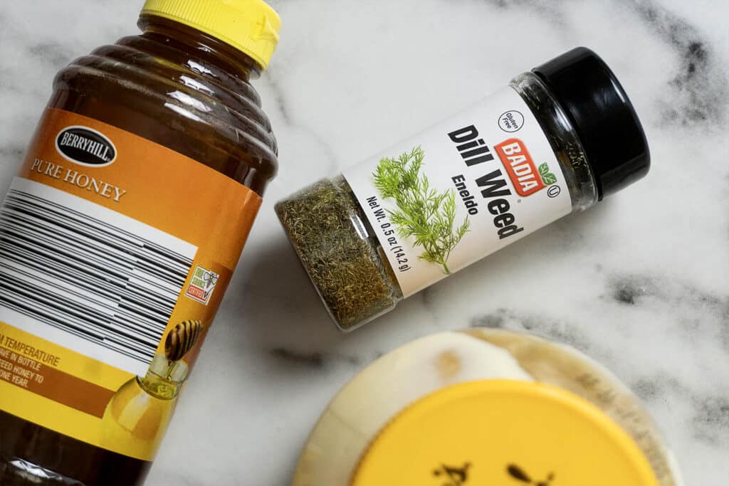 A bottle of Berryhill pure honey, a Badia dill weed spice container, and part of a round, yellow-capped jar are on a white marble surface.
